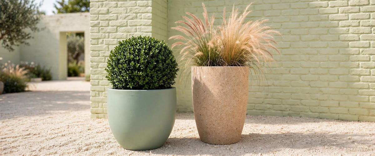 A smooth satin green fiberglass planter with clipped boxwood beside a taller stone-textured planter with ornamental grasses, on gravel in a lime-washed brick courtyard