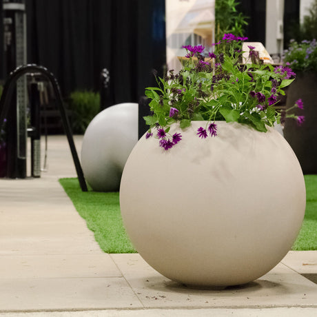 Orbo spherical Cosapots fiberglass planter in Sand Stone, shown planted with flowering perennials outdoors.
