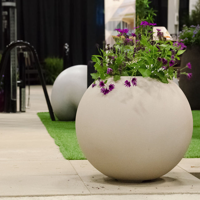 Orbo spherical Cosapots fiberglass planter in Sand Stone, shown planted with flowering perennials outdoors.