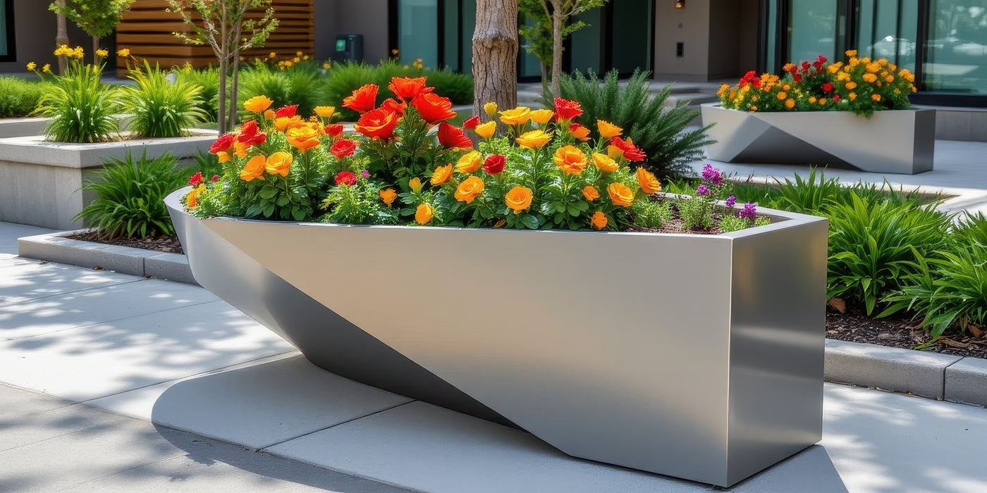 Angular brushed-metal planter filled with orange and red flowers on a modern stone plaza